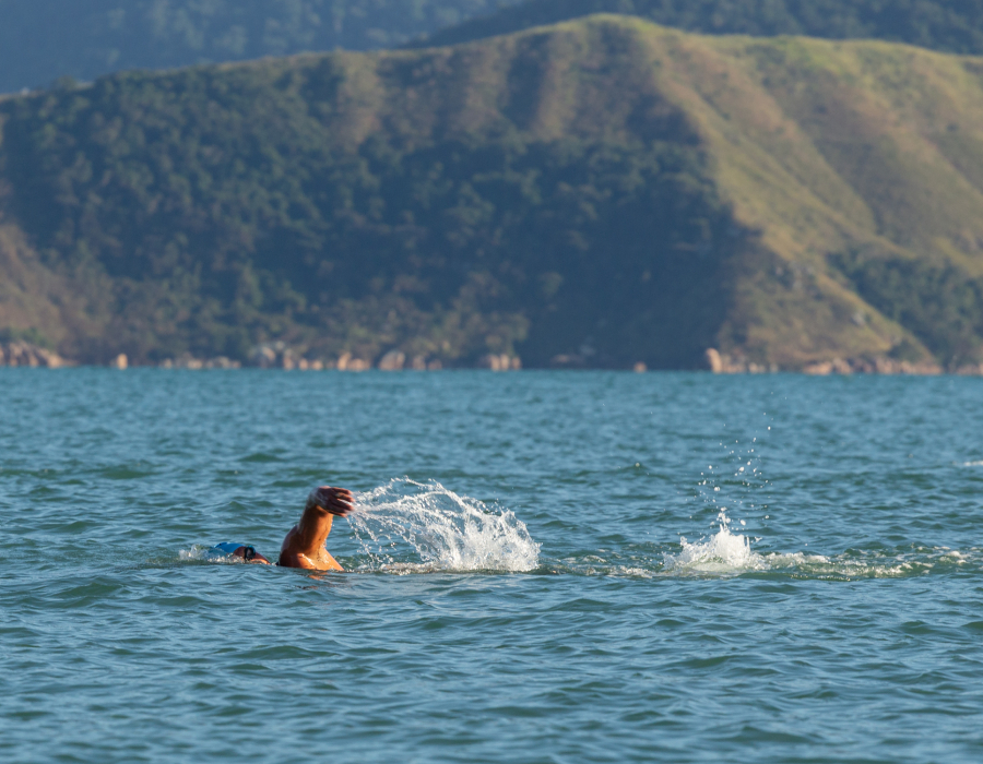Atleta nadando uma ultramaratona aquática, com água sendo levantada pelo braço no mar. Ao fundo, uma montanha